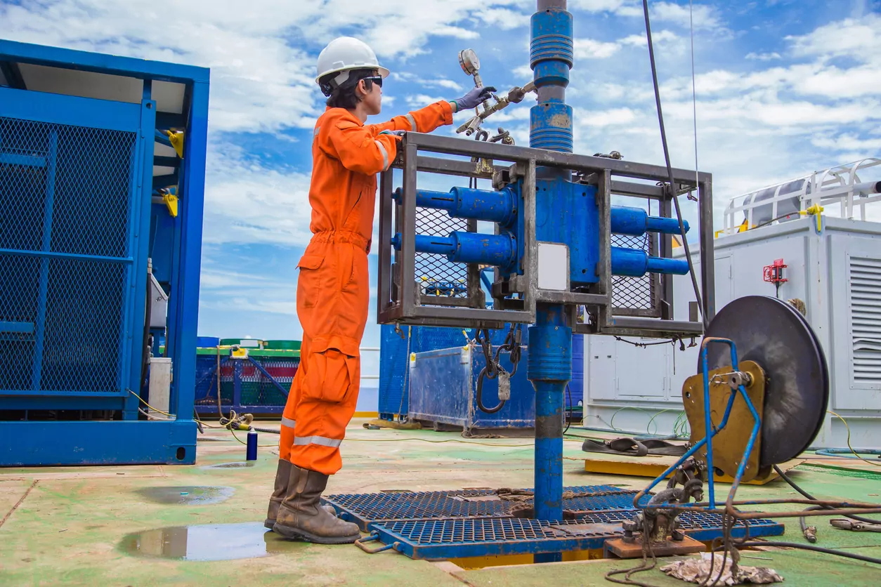Offshore wellsite operator in orange coveralls working at a wellhead with pressure control equipment and a wireline reel on deck